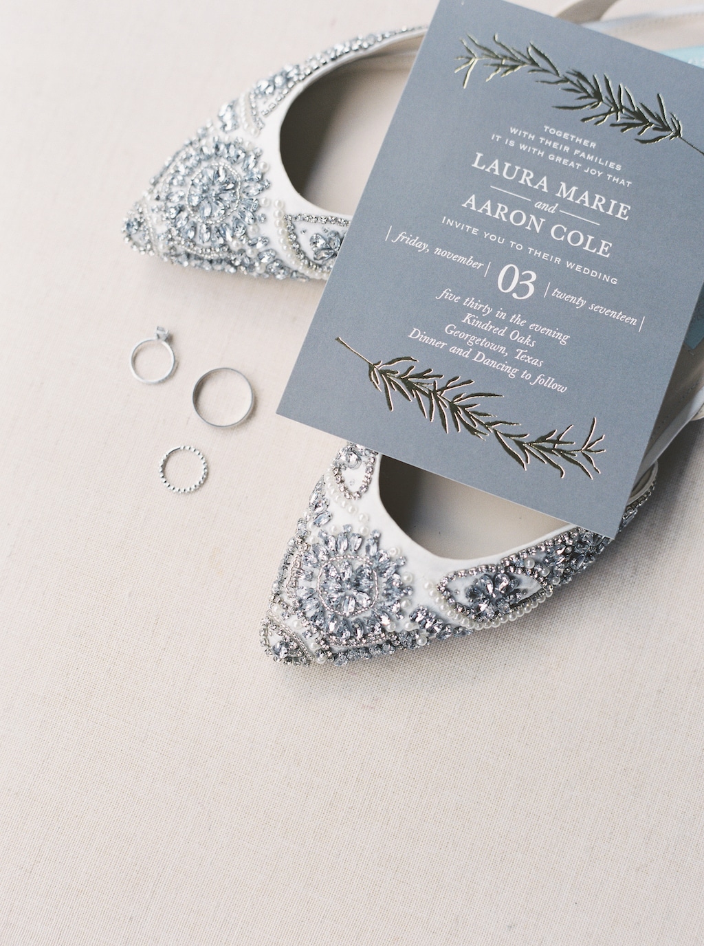 An invitation to a wedding at Kindred Oaks in Georgetown, Texas, rests on top of a pair of glamorous bridal shoes, next to the bride and groom's wedding rings.