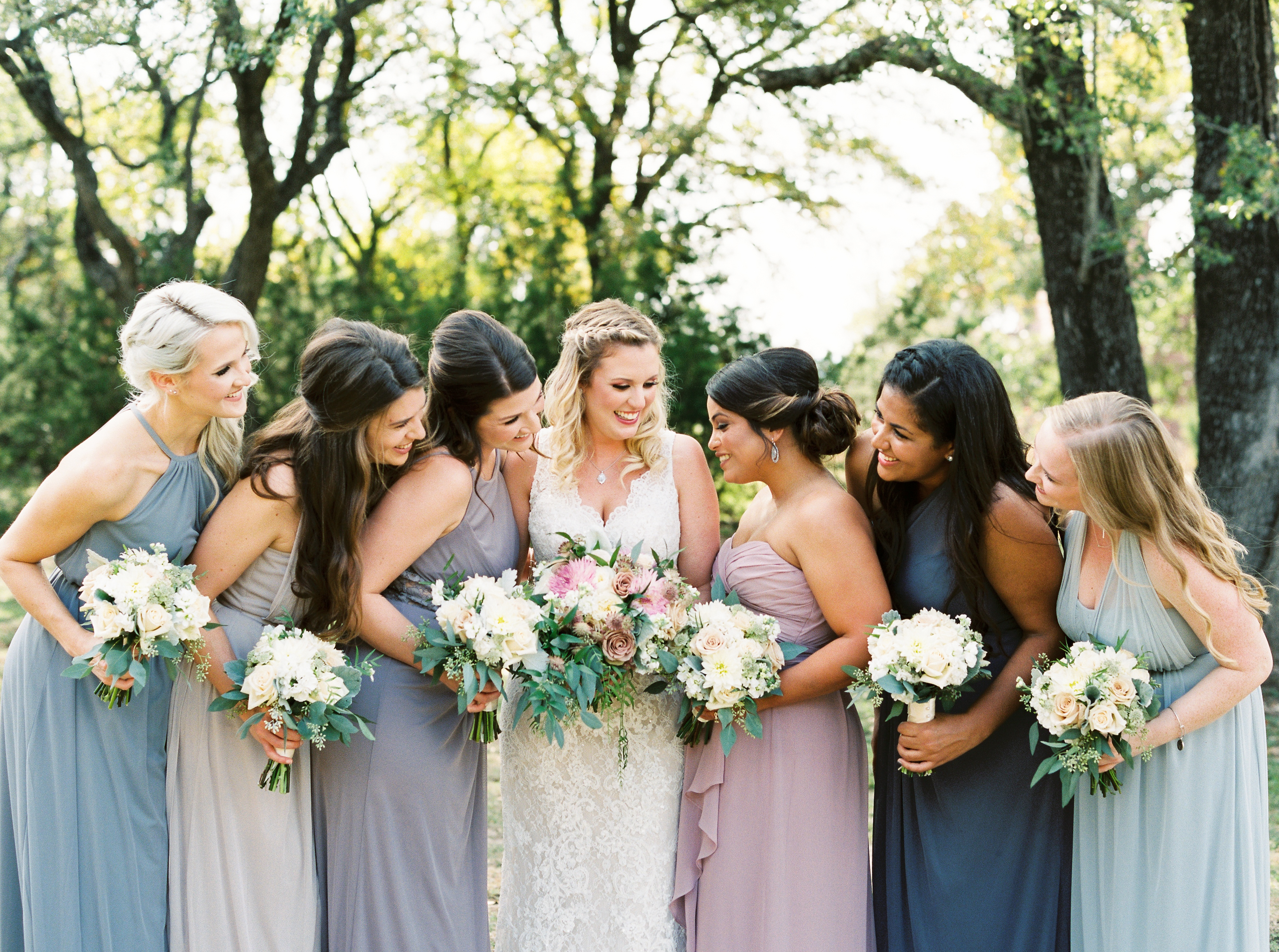 A bride and her six bridesmaids, wearing different shades of blue, stand smiling together on the grounds of Georgetown, Texas wedding venue, Kindred Oaks.