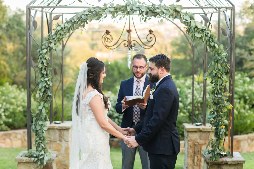 A bride and groom hold hands during their wedding ceremony in Georgetown, Texas as an officiant reads from the bible. Photo by Tank Goodness Photography.