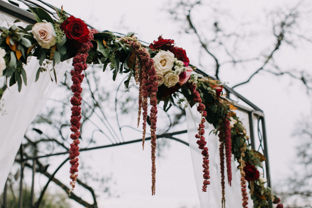 An iron arbor is decorated with red and white florals at an outdoor wedding venue in Georgetown, Texas. Photo by Tim Waters Photography.