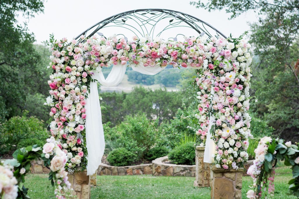 A large wrought iron wedding arbor is covered in layers of pink, white, and purple roses. Floral design by ZuZu's Petals of Austin.