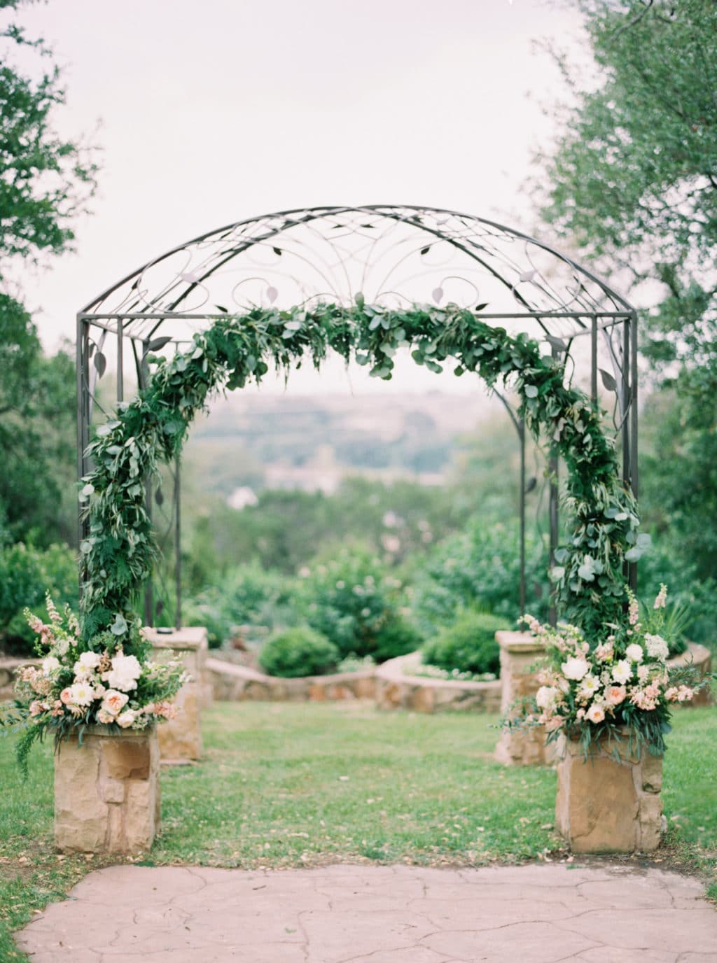 An intricate iron wedding arbor is decorated with an arch of greenery and muted florals at Kindred Oaks in Georgetown, Texas. Photo by Jenna McElroy Photography.